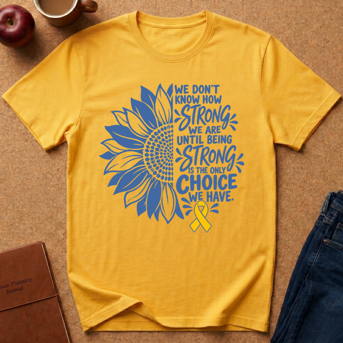 A yellow short-sleeve T-shirt laid flat on a brown cork-like surface, featuring a large blue sunflower graphic and the text 'We don't know how strong we are until being strong is the only choice we have,' plus a yellow ribbon. A red apple, coffee cup, notebook, and jeans are partially visible around it as styling props.