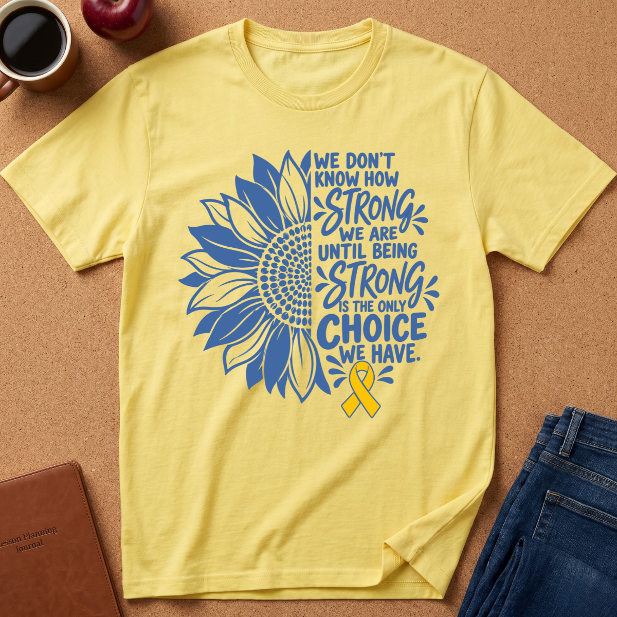 A pale yellow short-sleeve T-shirt laid flat on a cork surface, featuring a large blue sunflower graphic and the text 'We don't know how strong we are until being strong is the only choice we have,' plus a yellow awareness ribbon. A coffee cup, apple, notebook, and jeans are partially visible around it as styling props.