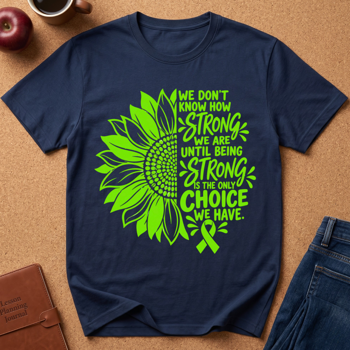 A navy blue short-sleeve t-shirt with a bright green sunflower graphic and the text 'WE DON'T KNOW HOW STRONG WE ARE UNTIL BEING STRONG IS THE ONLY CHOICE WE HAVE.' and a green ribbon, shown in a flat-lay arrangement on a cork background with jeans, a book, and a coffee cup nearby.