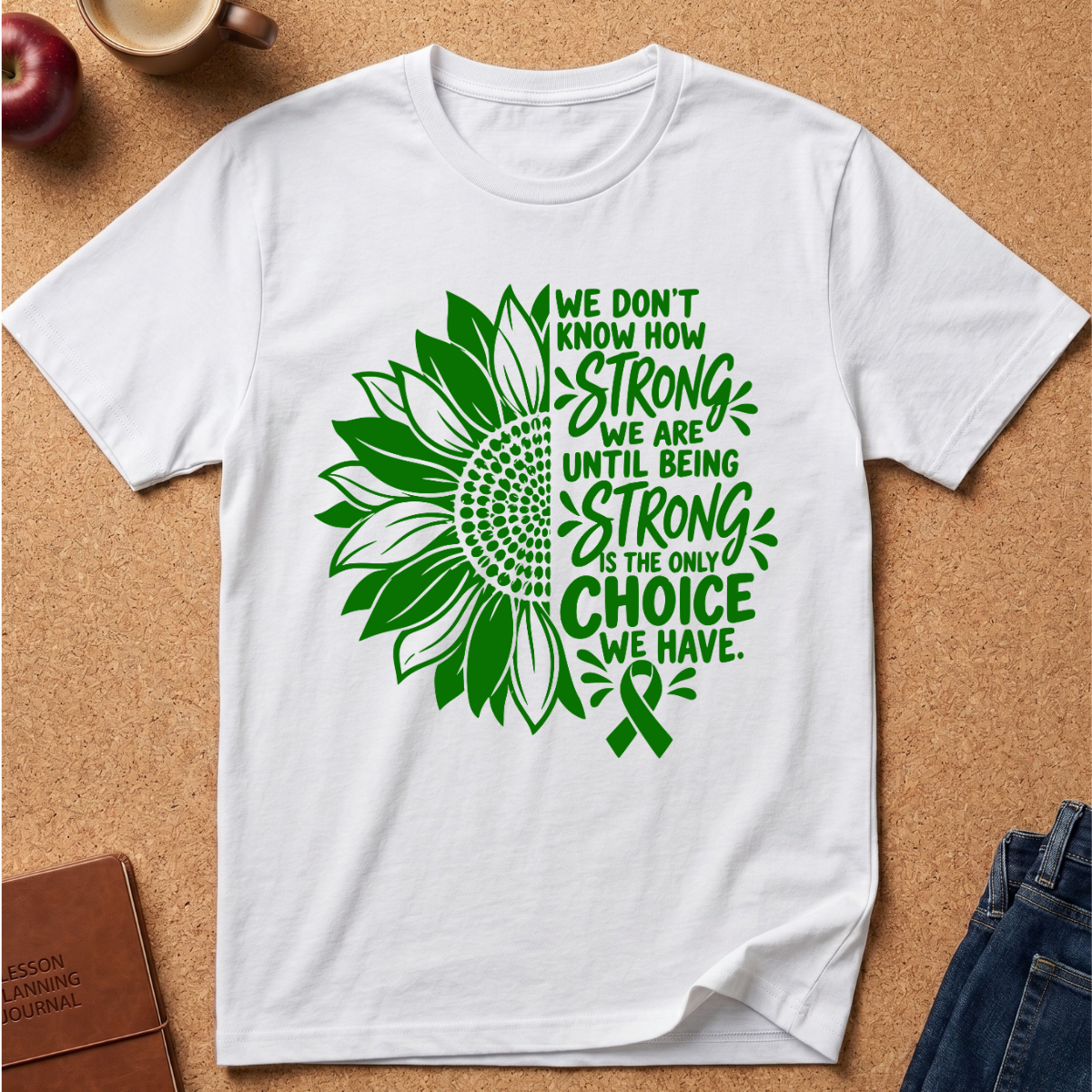 A white short-sleeve T-shirt laid flat on a cork surface, featuring a green sunflower graphic and the text 'WE DON'T KNOW HOW STRONG WE ARE UNTIL BEING STRONG IS THE ONLY CHOICE WE HAVE.' alongside a green awareness ribbon.