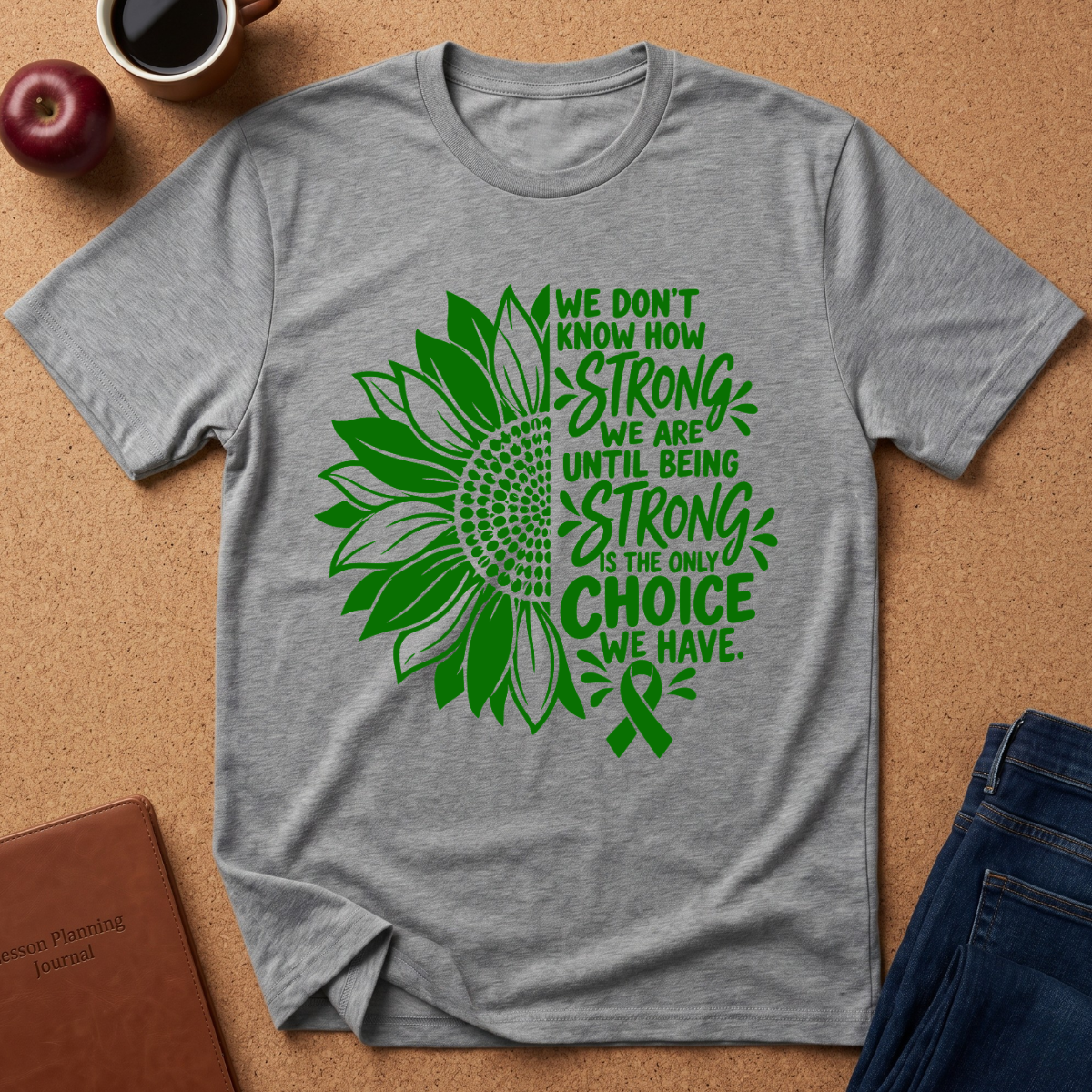 A heather gray short-sleeve T-shirt laid flat on a cork surface, featuring a green sunflower and ribbon graphic with the text 'WE DON'T KNOW HOW STRONG WE ARE UNTIL BEING STRONG IS THE ONLY CHOICE WE HAVE.' A red apple, coffee cup, notebook, and folded jeans are placed around it as styling props.