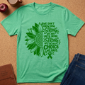 A mint green short-sleeve t-shirt laid flat on a cork background, featuring a large green sunflower and ribbon graphic with the text 'WE DON'T KNOW HOW STRONG WE ARE UNTIL BEING STRONG IS THE ONLY CHOICE WE HAVE.' Props including an apple, coffee cup, notebook, and jeans are partially visible around it.