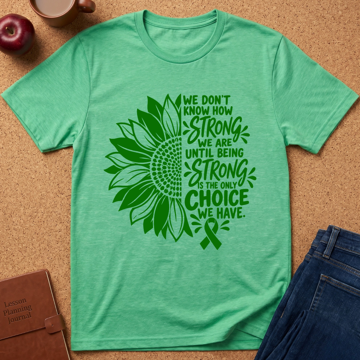 A mint green short-sleeve t-shirt laid flat on a cork background, featuring a large green sunflower and ribbon graphic with the text 'WE DON'T KNOW HOW STRONG WE ARE UNTIL BEING STRONG IS THE ONLY CHOICE WE HAVE.' Props including an apple, coffee cup, notebook, and jeans are partially visible around it.