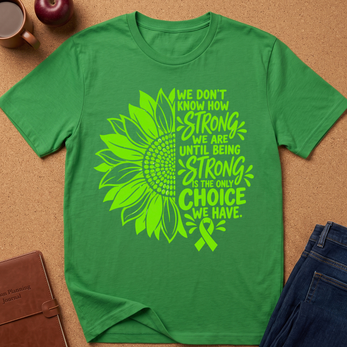 A green short-sleeve T-shirt laid flat on a cork surface, featuring a large neon green sunflower graphic and the text 'We don't know how strong we are until being strong is the only choice we have.' with a ribbon motif.
