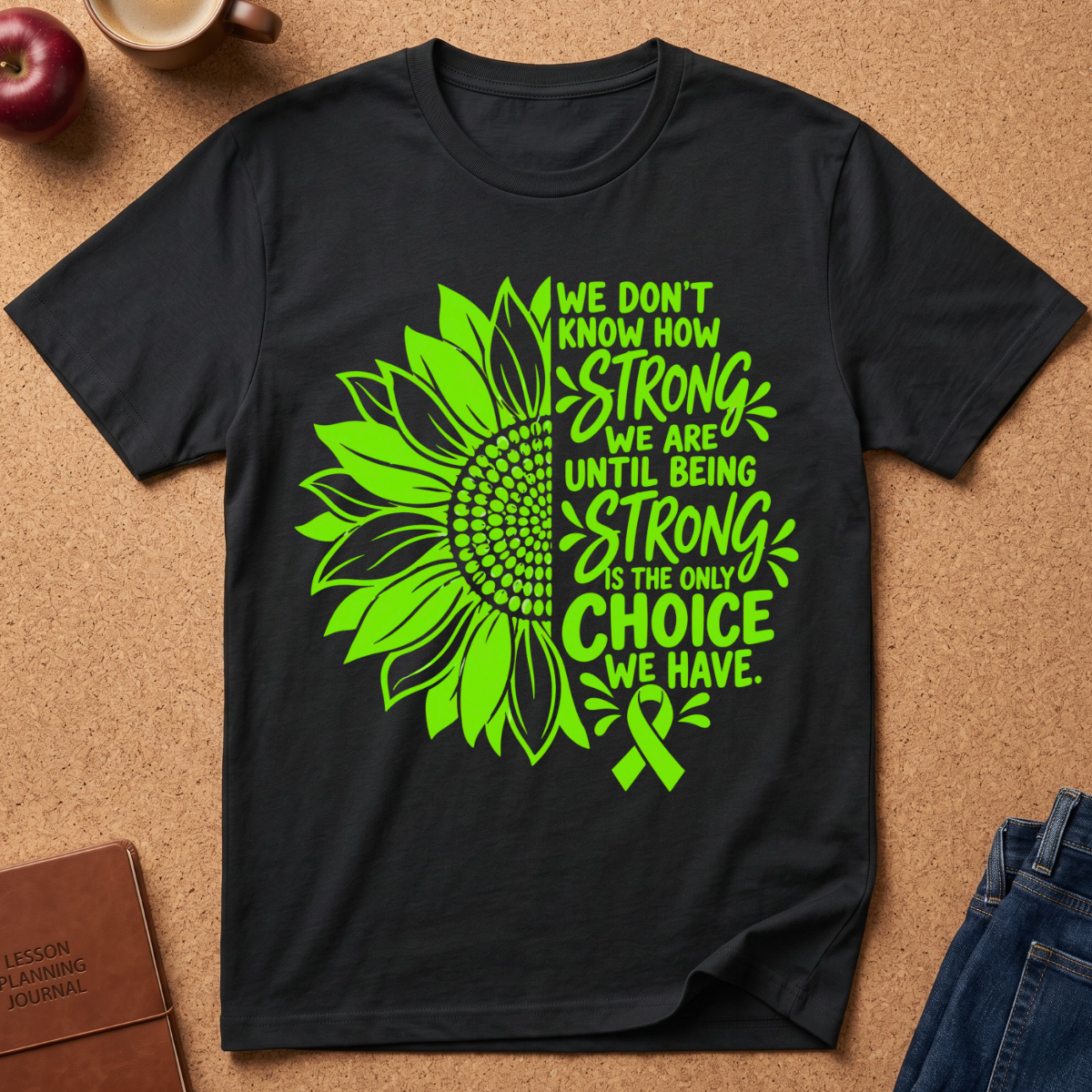 A black short-sleeve graphic T-shirt laid flat on a cork surface, featuring a large neon green sunflower and awareness ribbon design with the text 'We don't know how strong we are until being strong is the only choice we have.' A brown journal, apple, mug, and jeans are partially visible around the shirt as styled props.