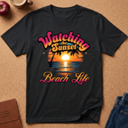 A black short-sleeve graphic t-shirt laid flat on a brown cork-like surface, featuring a retro sunset beach scene with palm trees and the text 'Watching the Sunset Beach Life' in pink, orange, and yellow lettering. Partial props including an apple, a mug, a journal, and jeans are visible around the shirt.