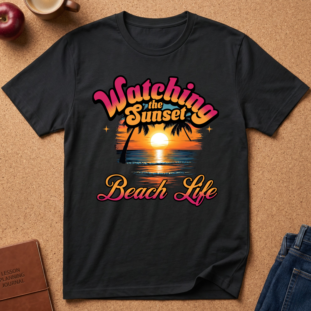 A black short-sleeve graphic t-shirt laid flat on a brown cork-like surface, featuring a retro sunset beach scene with palm trees and the text 'Watching the Sunset Beach Life' in pink, orange, and yellow lettering. Partial props including an apple, a mug, a journal, and jeans are visible around the shirt.