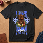 A black short-sleeve graphic T-shirt laid flat on a cork surface, featuring a colorful tiki mask, blue flames, flowers, and the text 'SUMMER VACATION TIKI HOUR.' Partial props around it include an apple, a coffee cup, a journal, and jeans.