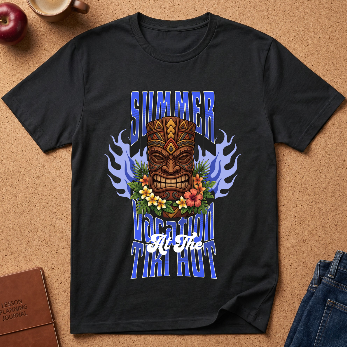 A black short-sleeve graphic T-shirt laid flat on a cork surface, featuring a colorful tiki mask, blue flames, flowers, and the text 'SUMMER VACATION TIKI HOUR.' Partial props around it include an apple, a coffee cup, a journal, and jeans.