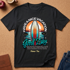A black short-sleeve graphic T-shirt laid flat on a cork surface, featuring a retro surfboard and sunset design with the text 'HAVE A NICE VACATION Good Day' and a summer quote. Partial props including an apple, mug, journal, and jeans are visible around the shirt.