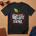A black short-sleeve graphic T-shirt laid flat on a cork surface, featuring a red apple outline with green leaves and the phrase 'TOO BUSY BEING A BRILLIANT TEACHER!' in white text. Surrounding props include a coffee cup, an apple, a journal, and folded jeans.