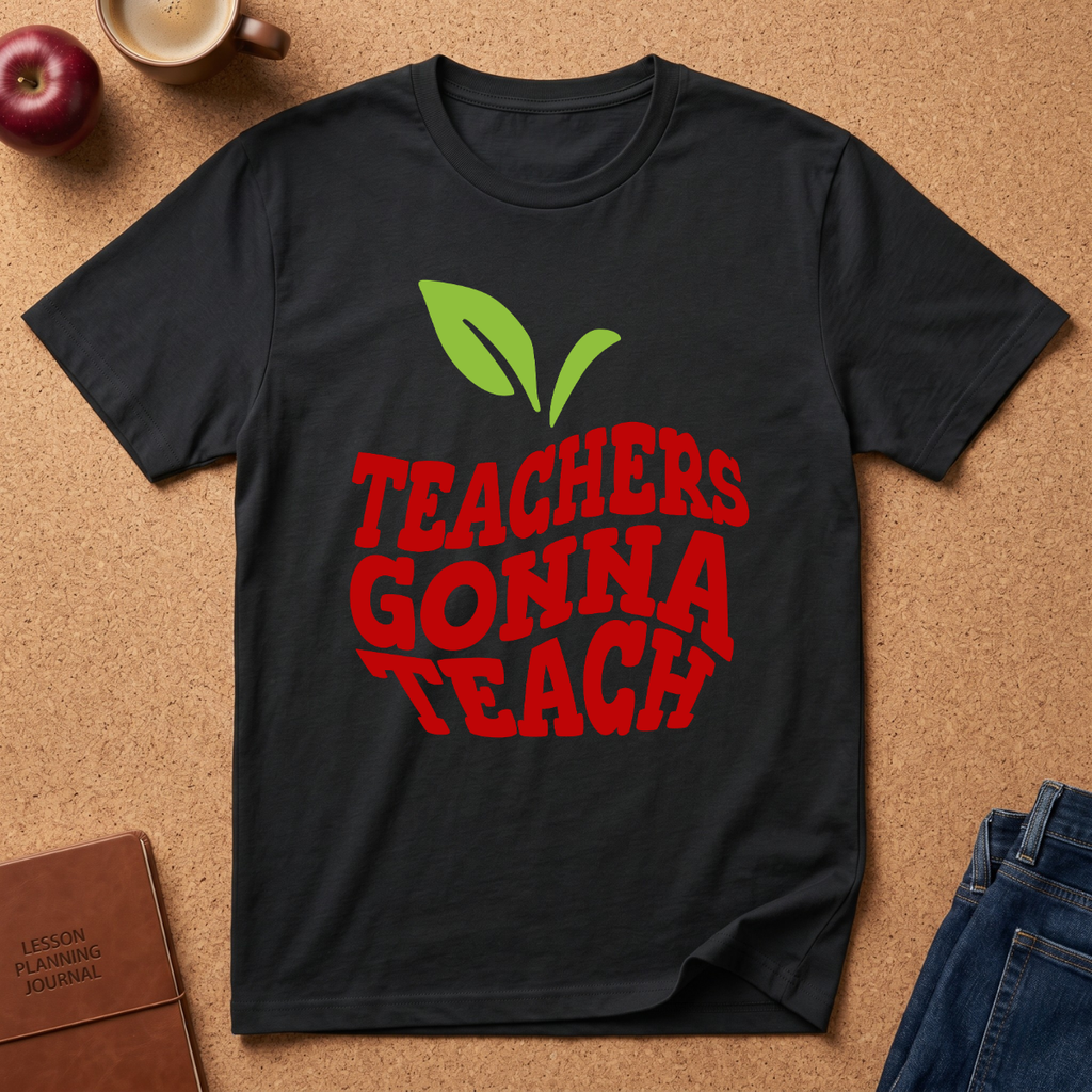 A black short-sleeve graphic T-shirt laid flat on a cork surface, featuring a red 'TEACHERS GONNA TEACH' design with green apple leaves above it. Surrounding props include an apple, a coffee cup, a journal, and folded blue jeans.