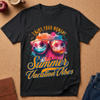 A black short-sleeve graphic T-shirt laid flat on a cork background, featuring colorful text reading 'Enjoy Your Moment Summer Vacation Vibes' and a pair of sunglasses with palm tree reflections. Partial props around the shirt include an apple, a cup, a journal, and jeans.