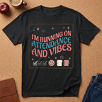 Black t-shirt with colorful text and graphics on a brown surface with an apple, coffee, and jeans.   I'm running on Attendance and Vibes.