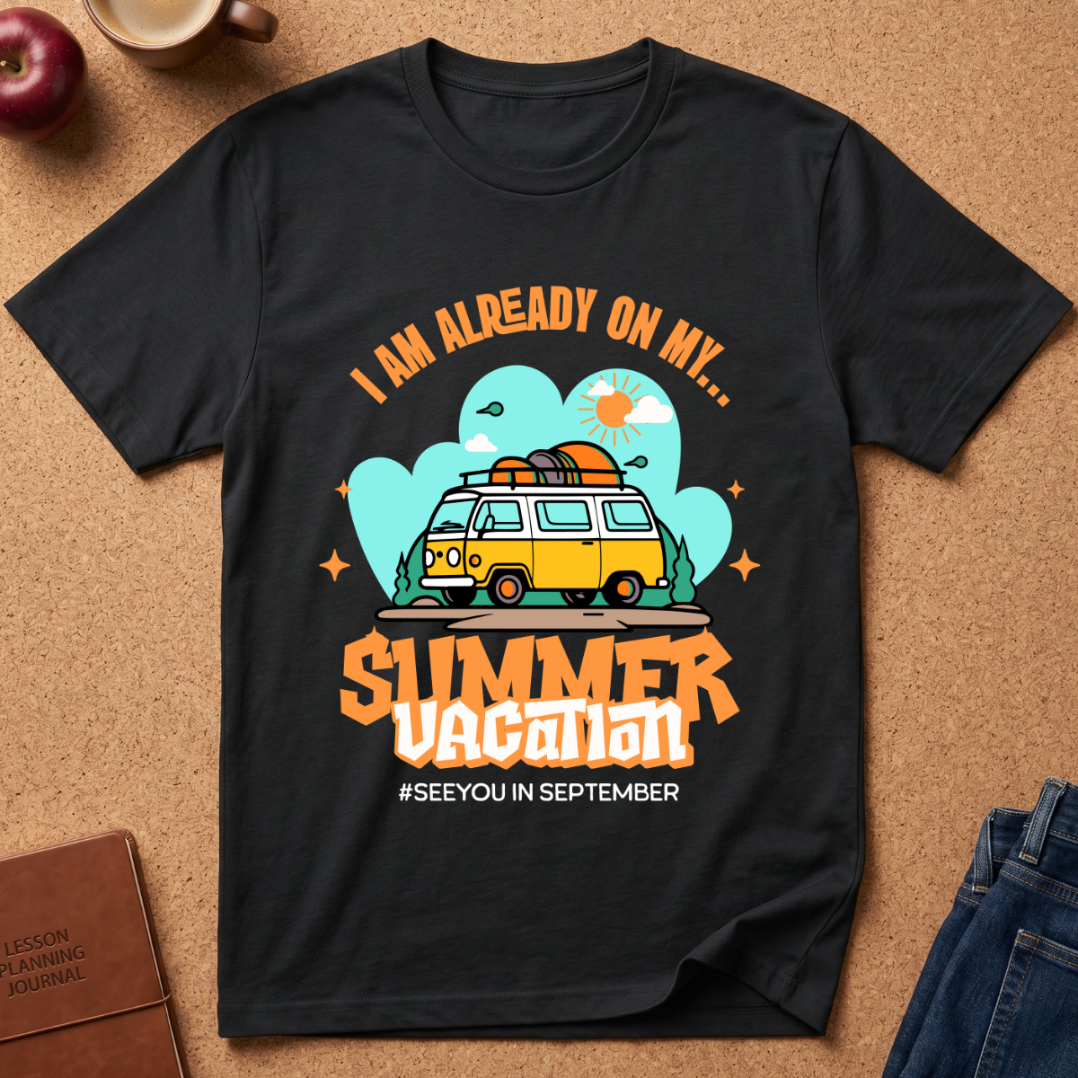 A black  graphic t-shirt laid flat on a cork surface, surrounded by lifestyle props including a brown leather journal, an apple, a coffee cup, and folded blue jeans. The t-shirt features a retro-style graphic of a yellow and white van with a surfboard on its roof, set against a light blue cloud background with the text 'I AM ALREADY ON MY sUMMER VACATION. #SEEYOUINSEPTEMBER