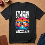 A black graphic t-shirt laid flat on a cork background, featuring the text 'I'M GIVING SUMMER VACATION VIBES' in white around a colorful retro sunset and sunglasses design.