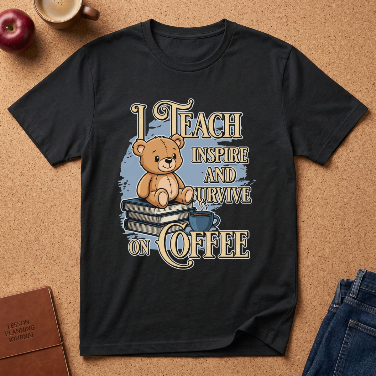 A black short-sleeve graphic T-shirt laid flat on a cork surface, featuring a teddy bear sitting on books with the text 'I TEACH INSPIRE AND SURVIVE ON COFFEE.' The shirt is styled with a coffee cup, apple, notebook, and jeans around it.