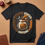 Black graphic t-shirt says "Current Status, Powered by Caffeine and Chaos" with coffee-themed graphic and text on a brown surface with coffee and jeans.