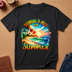 A black short-sleeve graphic T-shirt laid flat on a cork-like surface, featuring a colorful tropical beach scene with palm trees and the text 'SCHOOL'S OUT FOR SUMMER.' Partial props around the shirt include an apple, a mug, a journal, and jeans.