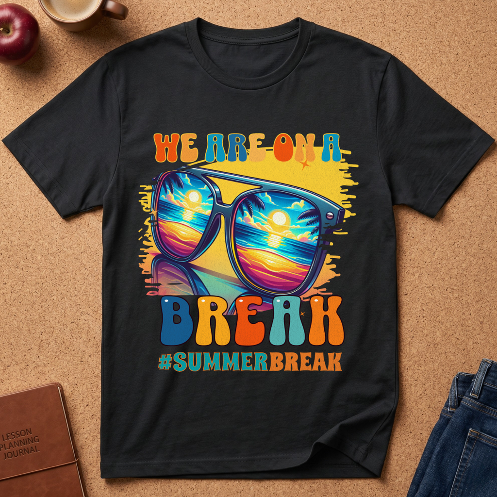 A black graphic t-shirt laid flat on a cork background, featuring a colorful 'WE ARE ON A SUMMER BREAK' design with sunglasses reflecting a sunset scene. Styled flat-lay props include an apple, a coffee cup, a journal, and jeans.