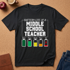 Black t-shirt with 'Battery Life of a Elementary School Teacher' graphic on a brown surface with school supplies.