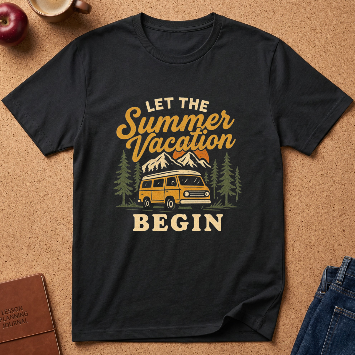 A black short-sleeve graphic T-shirt laid flat on a cork surface, featuring a retro-style summer vacation design with a yellow van, mountains, trees, and the text 'LET THE Summer Vacation BEGIN.' Partial props around it include an apple, a mug, a journal, and folded blue jeans.