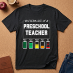 Black t-shirt with 'Battery Life of a Preschool Teacher' graphic on a brown surface with school supplies.