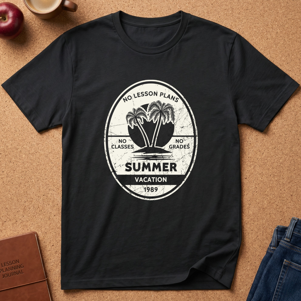 A black short-sleeve graphic T-shirt laid flat on a cork-like surface, featuring a distressed white circular print with palm trees and the words 'No Lesson Plans,' 'Summer Vacation,' and '1989.' A brown journal, apple, mug, and blue jeans are partially visible around it as styling props.