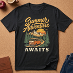 A black short-sleeve graphic T-shirt laid flat on a cork-like surface, featuring a retro-style 'Summer Adventure AWAITS' print with a mountain, trees, campervan, and sunset. Partial props including an apple, mug, journal, and jeans are visible around the shirt.