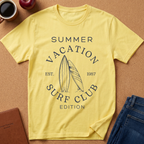 A pale yellow graphic T-shirt laid flat on a cork surface, surrounded by styled props including a coffee mug, apple, notebook, and folded jeans. The shirt has a dark navy printed design reading “SUMMER VACATION SURF CLUB EST.