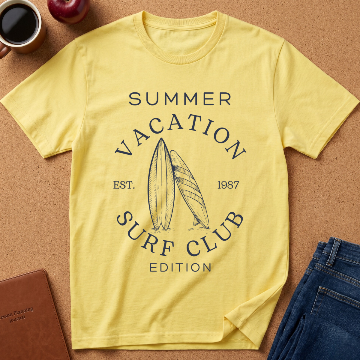 A pale yellow graphic T-shirt laid flat on a cork surface, surrounded by styled props including a coffee mug, apple, notebook, and folded jeans. The shirt has a dark navy printed design reading “SUMMER VACATION SURF CLUB EST.