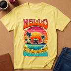 A pale yellow graphic T-shirt laid flat on a cork-like surface, surrounded by a coffee mug, apple, notebook, and folded jeans. The shirt has a large retro sunset beach graphic with palm trees, a van, birds, and the words “HELLO SUMMER” in bold multicolor lettering.