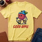 A pale yellow graphic T-shirt laid flat on a cork surface, featuring a cartoon apple character wearing a blue beanie and sneakers with the words 'GOOD APPLE' in red below. Surrounding props include a coffee cup, an apple, a notebook, and folded jeans.