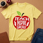 A yellow short-sleeve T-shirt laid flat on a cork surface, featuring a large red apple graphic with the words 'TEACH INSPIRE GROW' in white and a green leaf. A coffee mug, apple, notebook, and jeans are placed around it as styling props.