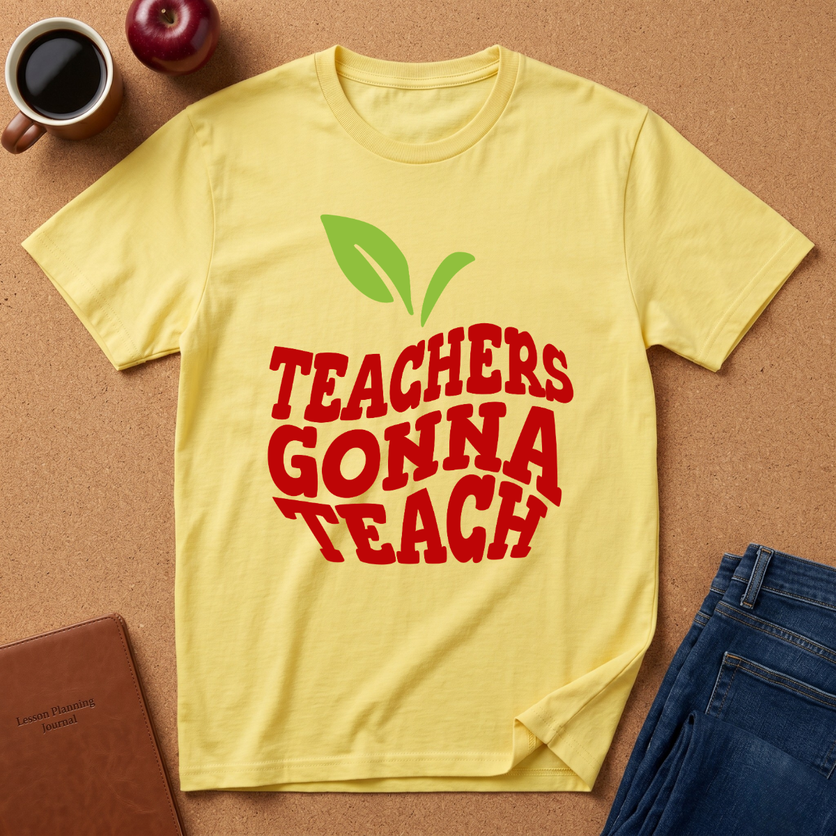 A yellow short-sleeve t-shirt with a red and green graphic reading 'TEACHERS GONNA TEACH' is laid flat on a cork background with a coffee mug, apple, notebook, and jeans as props.