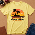 A pale yellow graphic T-shirt laid flat on a cork-like surface, surrounded by lifestyle props including a coffee mug, apple, notebook, and folded jeans. The shirt features a tropical sunset print with palm trees, birds, and the text 'Dreaming of Summer Vacation No More Classes.