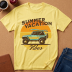 A pale yellow graphic T-shirt laid flat on a cork-like surface, with a retro sunset-style print showing a turquoise and black jeep, a surfboard, and the words “SUMMER VACATION Vibes.” Surrounding props include a coffee mug, apple, notebook, and folded blue jeans.