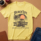 A pale yellow graphic T-shirt laid flat on a cork-like surface, surrounded by styled props including a coffee cup, apple, notebook, and blue jeans. The shirt features a retro beach-themed print with the words “BEACH LIFE,” a palm tree, a vintage camper van, and a sunset circle.
