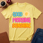 Yellow t-shirt with 'Good Morning Sunshine' text on a brown surface with a coffee cup and apple.