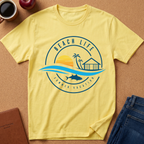 A pale yellow short-sleeve t-shirt laid flat on a cork-like surface, featuring a circular beach-themed graphic with the words 'BEACH LIFE' and 'SUMMER VACATION,' plus a sunset, palm trees, a beach house, waves, and a fish. Coffee cup, apple, notebook, and folded jeans are partially visible around it as styling props.