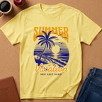 A pale yellow short-sleeve T-shirt laid flat on a cork-like surface, featuring a retro beach sunset graphic with a palm tree, blue waves, birds, and the words “SUMMER Vacation” and “SUN-SALT SAND.” Coffee cup, apple, notebook, and jeans are placed around it as styling props.