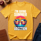A yellow graphic t-shirt laid flat on a brown textured surface, surrounded by lifestyle props including a red apple, a coffee mug, a brown notebook, and blue jeans. The t-shirt features a colorful retro graphic of sunglasses with a sunset landscape reflection and the text 'I'M GIVING SUMMER VACATION VIBES'.