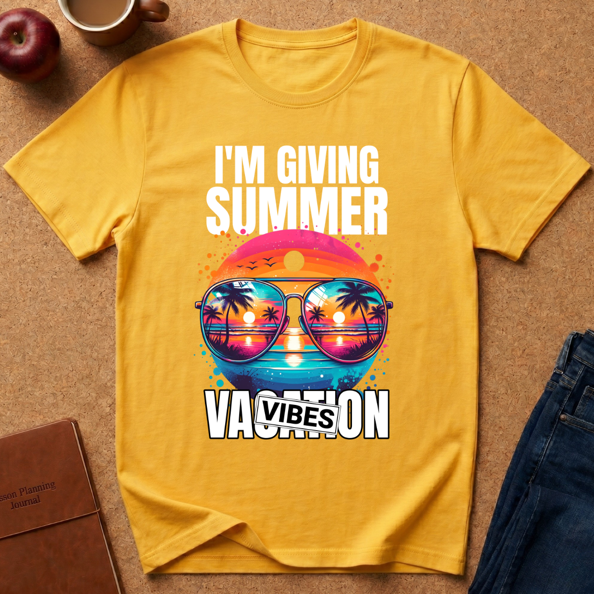 A yellow graphic t-shirt laid flat on a brown textured surface, surrounded by lifestyle props including a red apple, a coffee mug, a brown notebook, and blue jeans. The t-shirt features a colorful retro graphic of sunglasses with a sunset landscape reflection and the text 'I'M GIVING SUMMER VACATION VIBES'.