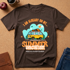 A dark brown  graphic t-shirt laid flat on a cork surface, surrounded by lifestyle props including a brown leather journal, an apple, a coffee cup, and folded blue jeans. The t-shirt features a retro-style graphic of a yellow and white van with a surfboard on its roof, set against a light blue cloud background with the text 'I AM ALREADY ON MY sUMMER VACATION. #SEEYOUINSEPTEMBER