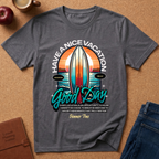 A dark gray graphic t-shirt laid flat on a cork background, surrounded by lifestyle props including a coffee cup, an apple, a journal, and jeans. The t-shirt features a colorful retro surfboard graphic with the text 'HAVE A NICE VACATION Good Day Summer Time'.