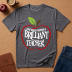 A dark gray heather t-shirt with a graphic of a red apple containing the text 'TOO BUSY BEING A BRILLIANT TEACHER!' in white. The shirt is displayed in a flat-lay arrangement on a cork background with a red apple, a coffee mug, a journal, and blue jeans.