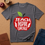 A dark gray heather t-shirt laid flat on a cork background, featuring a red apple graphic with the words 'TEACH INSPIRE GROW' in white. The shirt is styled with a brown notebook, an apple, a coffee mug, and blue jeans.