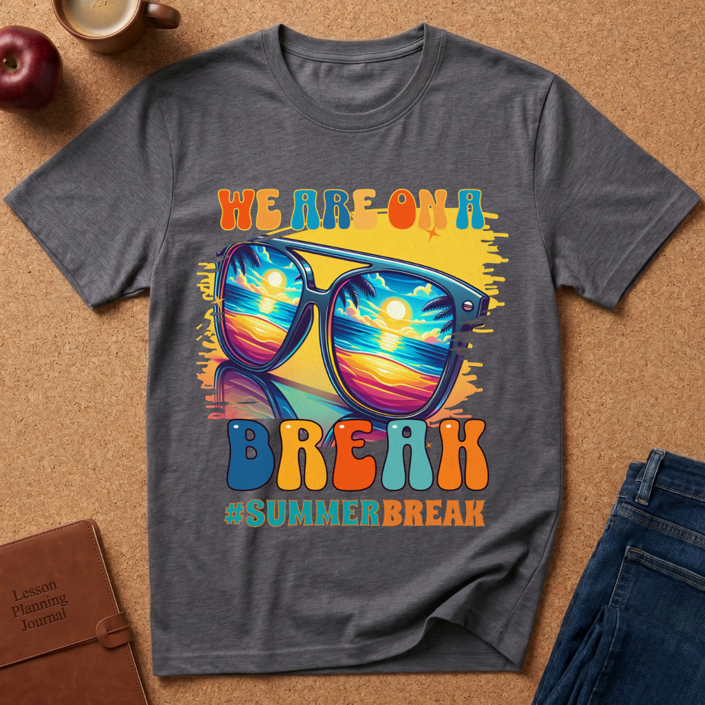 A dark gray graphic t-shirt laid flat on a cork background, featuring a colorful 'WE ARE ON A SUMMER BREAK' design with sunglasses reflecting a sunset scene. Styled flat-lay props include an apple, a coffee cup, a brown journal, and blue jeans.