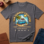 A dark gray graphic t-shirt laid flat on a cork background, featuring a retro-style illustration of a blue van on a beach with the text 'VAN SUMMER VACATION' and 'DRIVE INTO SUMMER WITH STYLE.' Styled props around it include an apple, a coffee cup, a brown journal, and blue jeans.