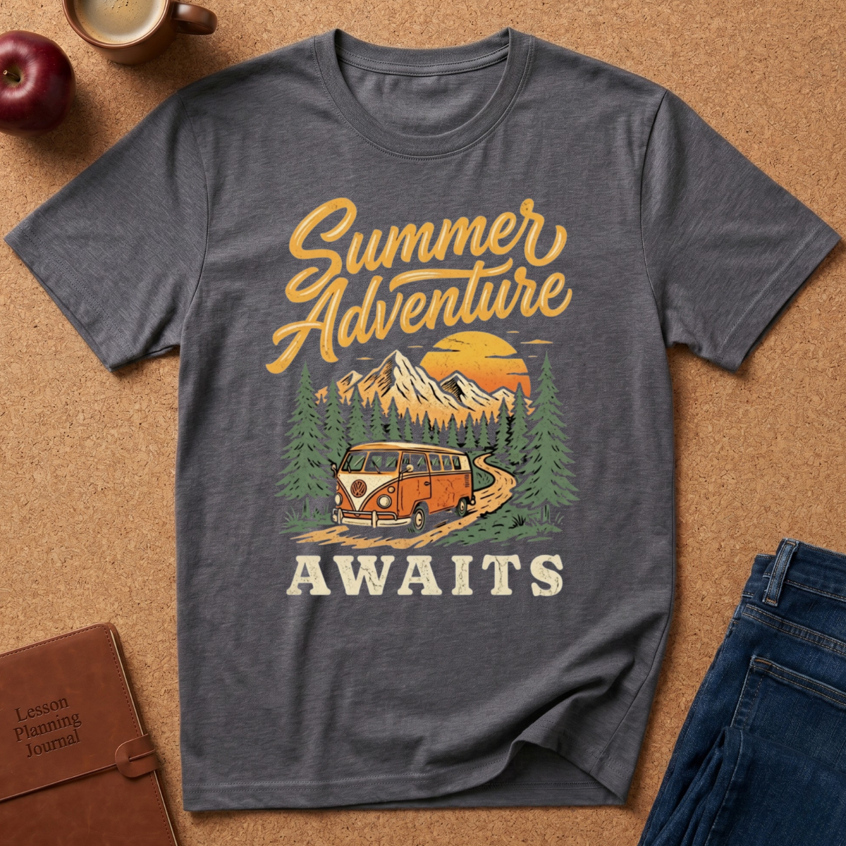 A heather dark gray graphic t-shirt laid flat on a cork background, featuring a retro-style 'Summer Adventure AWAITS' print with a mountain, pine trees, a campervan, and an orange sunset. Styled flat-lay props include an apple, a coffee cup, a journal, and blue jeans.